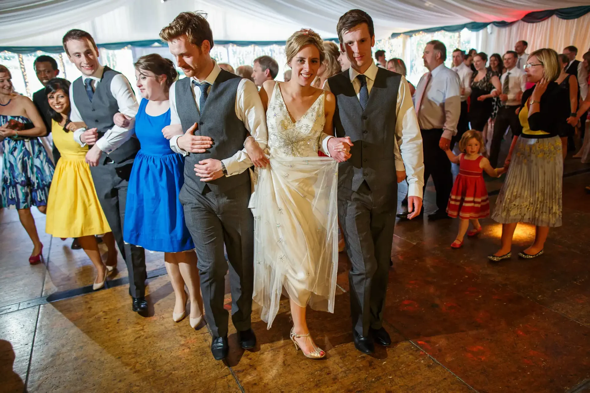 A bride and groom walk arm-in-arm flanked by friends in a festive wedding reception, with guests dancing around them.