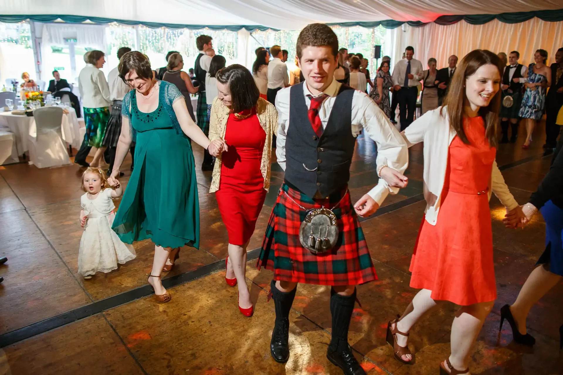 Group of people dancing at a wedding reception, including a man in a kilt and a little girl in a white dress.