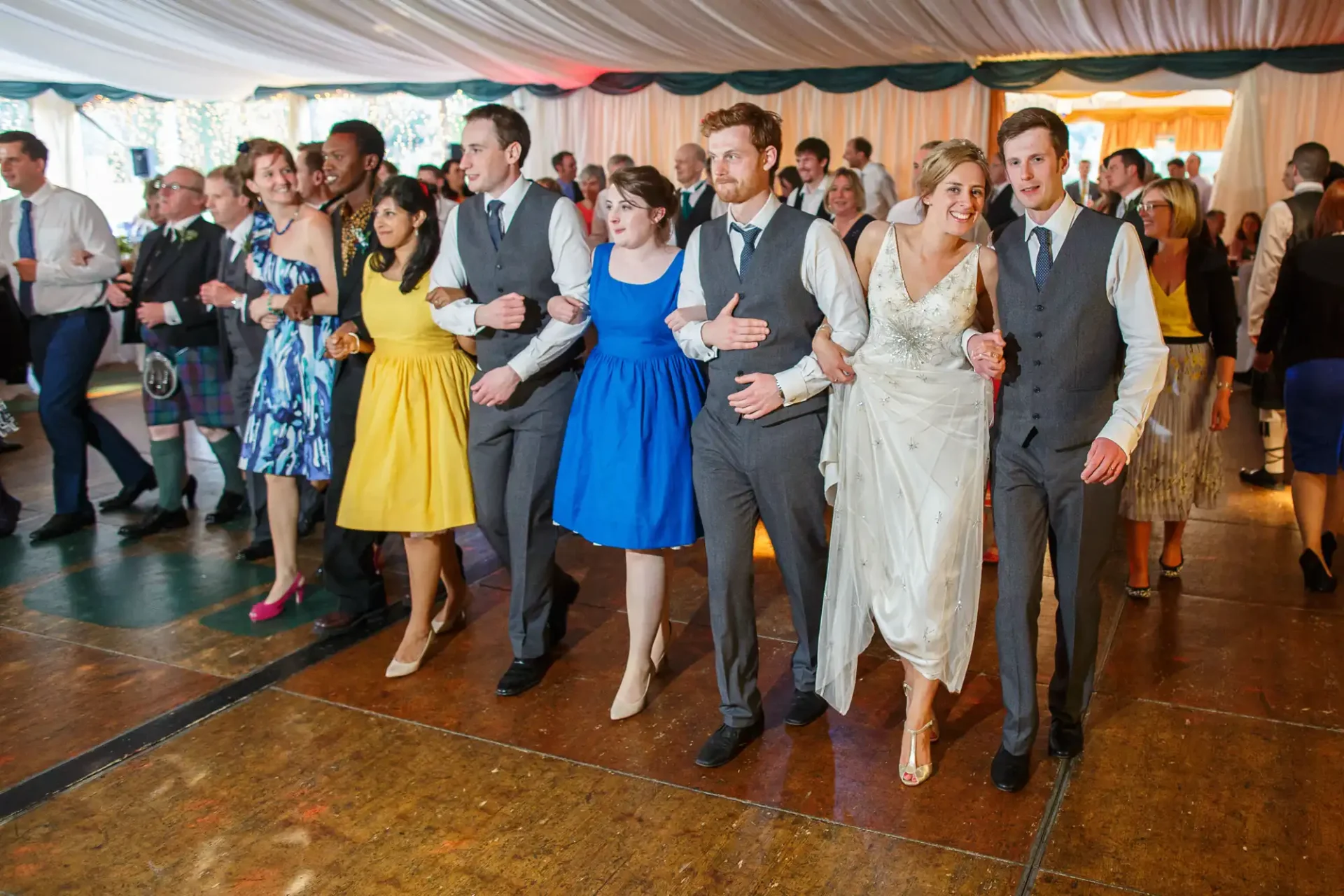 A joyful group of people, including a bride and groom, holding hands and dancing in a line at a wedding reception under a tent.