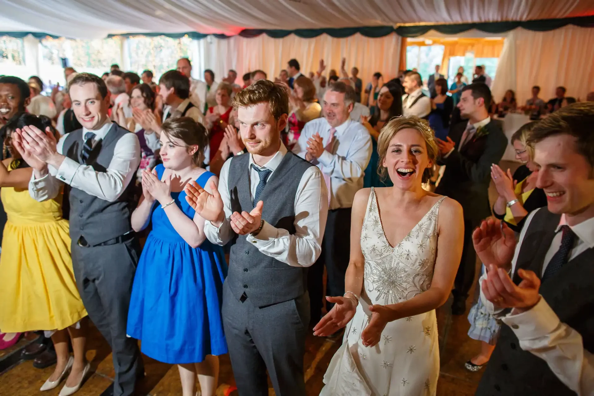 Wedding guests clapping and dancing in a lively reception tent, featuring a joyful bride and groom at the center.