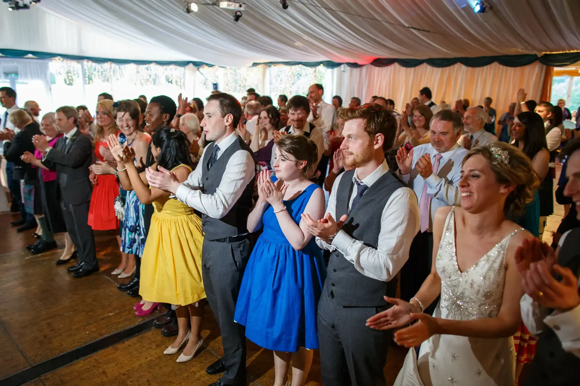A group of elegantly dressed people clapping and smiling at a wedding reception inside a tent.