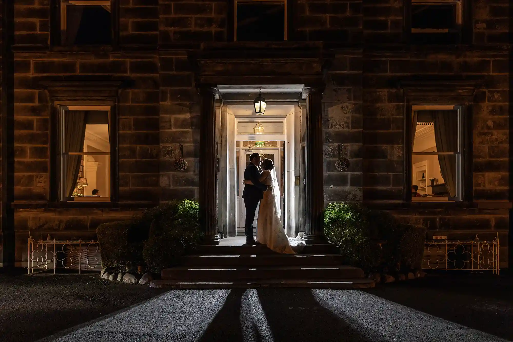 A newlywed couple embracing at the illuminated entrance of a stone building at night.