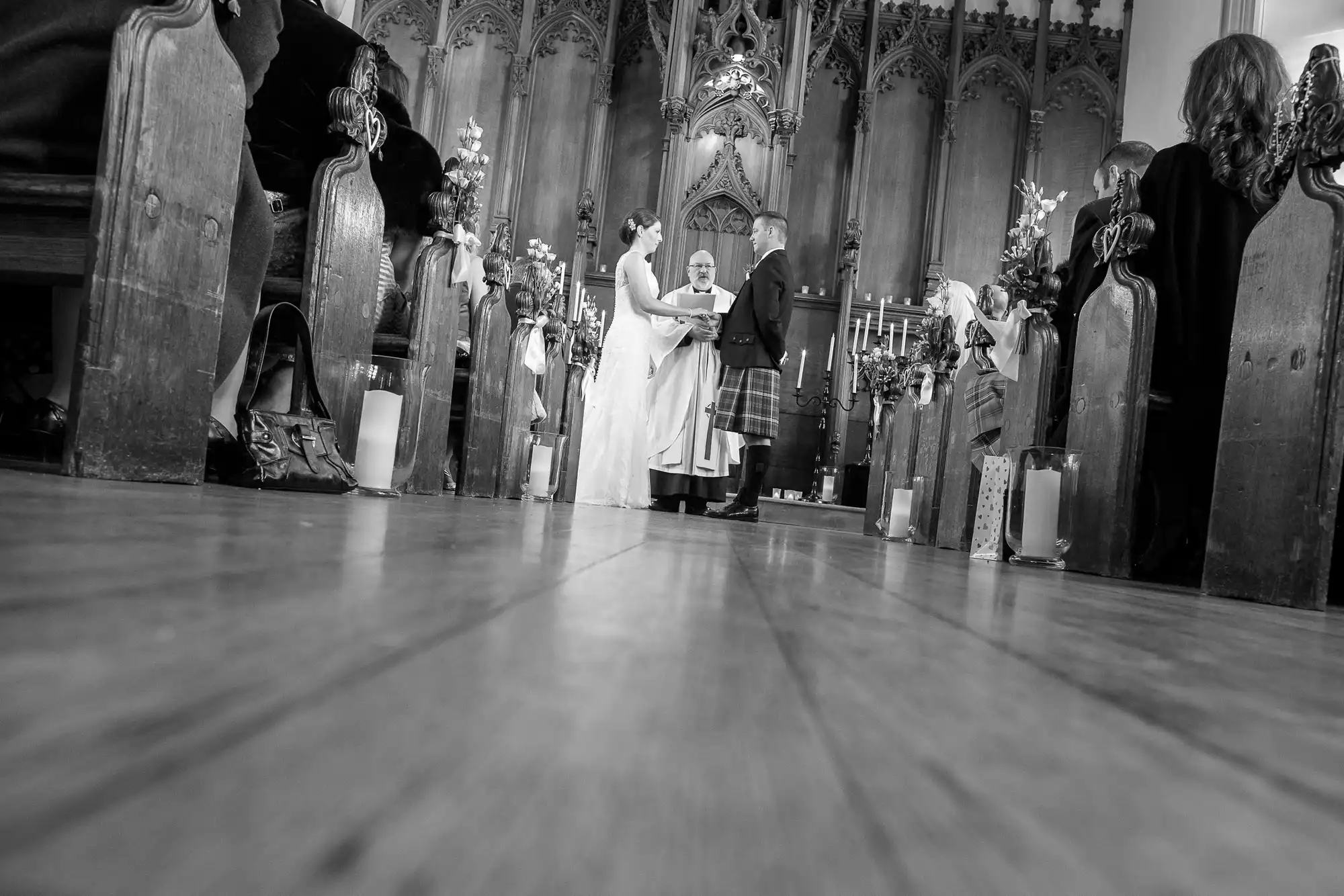 A bride and groom standing at the altar with a priest during a wedding ceremony, viewed from a low angle between wooden pews in a church.