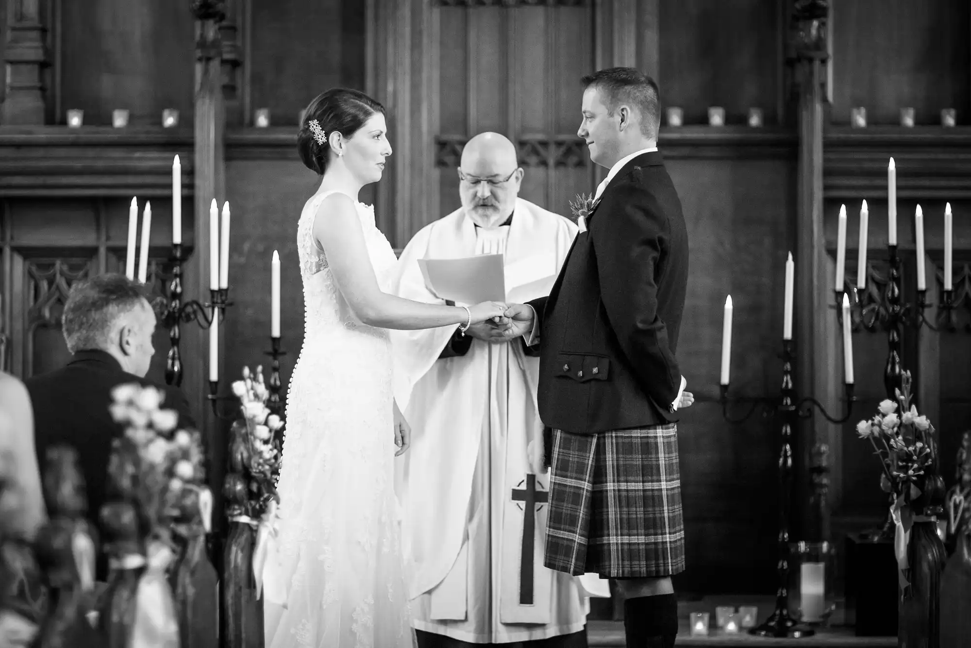 A bride and groom holding hands during their wedding ceremony, officiated by a priest in a church with lit candles.