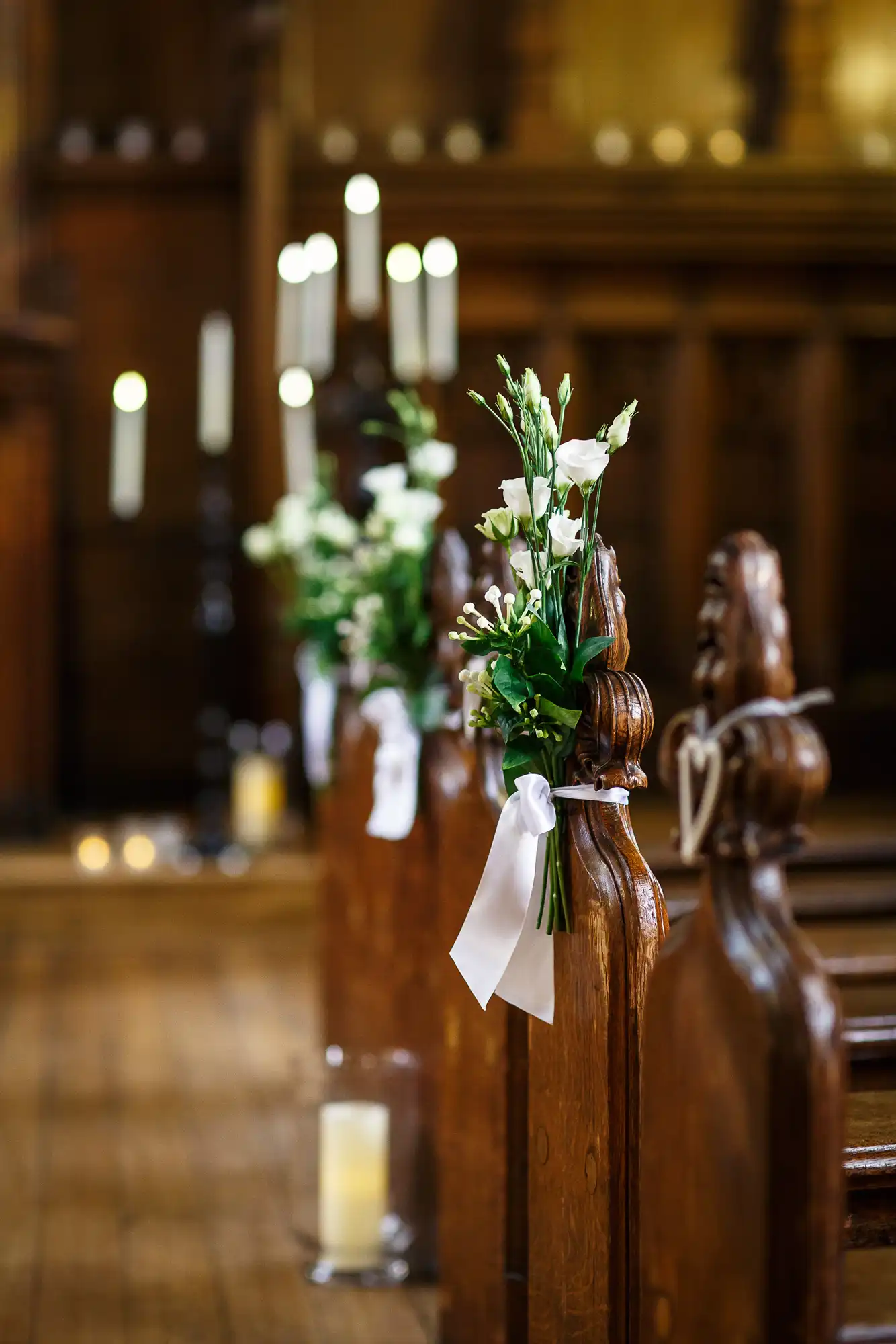 Wooden church pews decorated with white flowers and ribbons, candles in the background, in a dimly lit church setting.