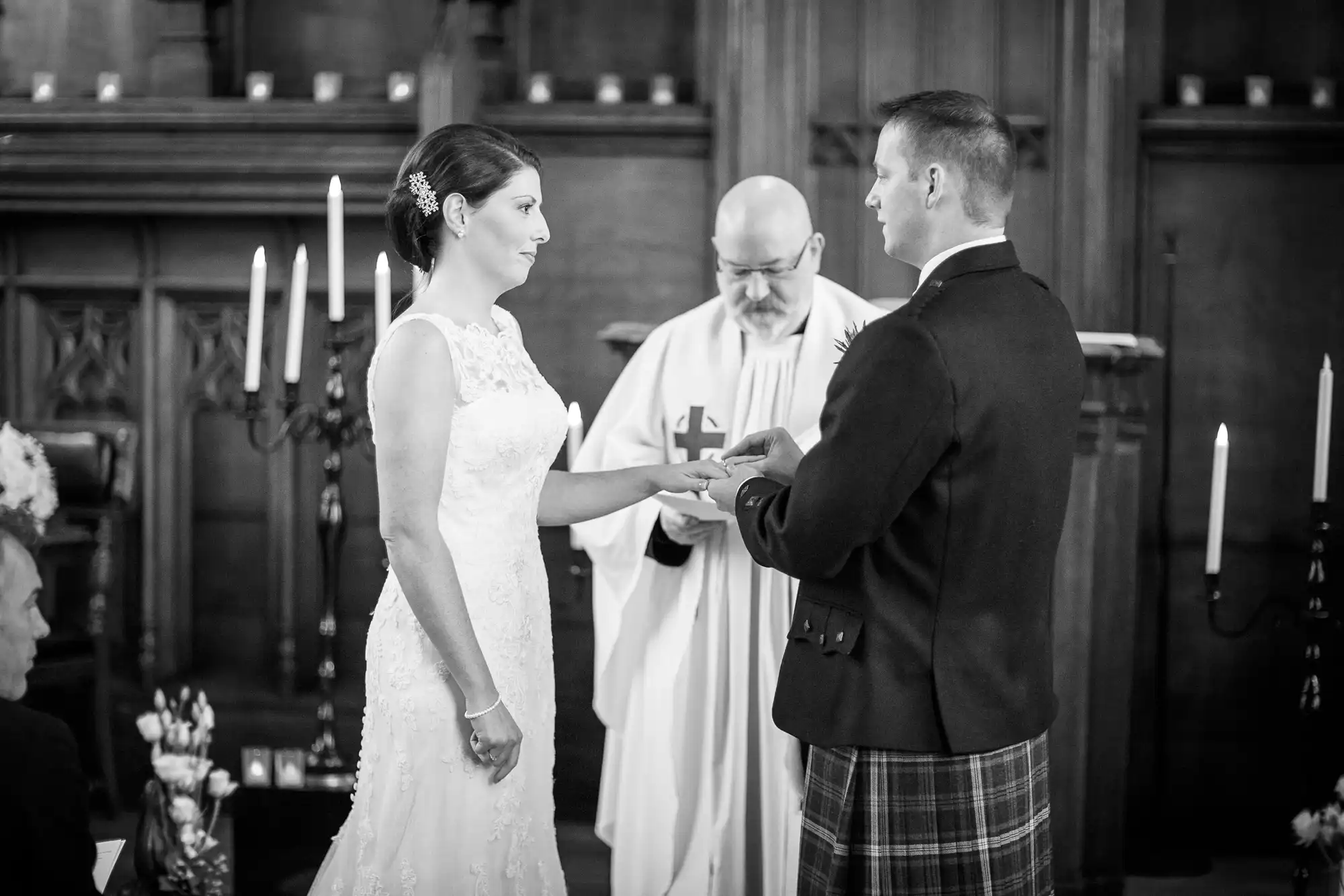 A bride in a white dress and a groom in a kilt exchange rings during a wedding ceremony in a church, officiated by a priest.