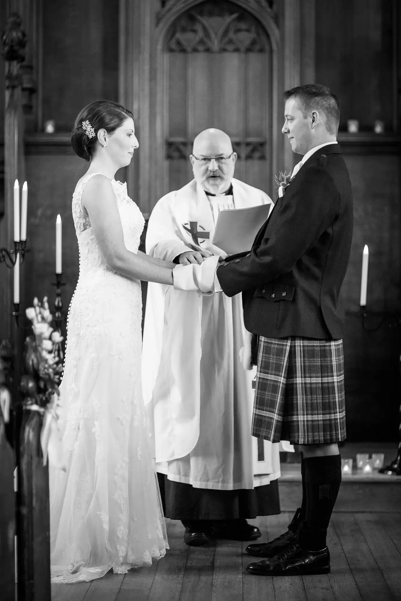 A bride and groom holding hands during a wedding ceremony, officiated by a priest in a church, captured in black and white.