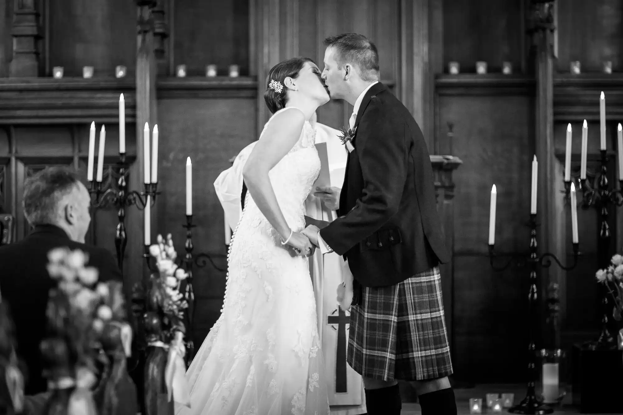 A bride in a white dress and a groom in a kilt share a kiss at their wedding inside a church with candlelit ambiance.