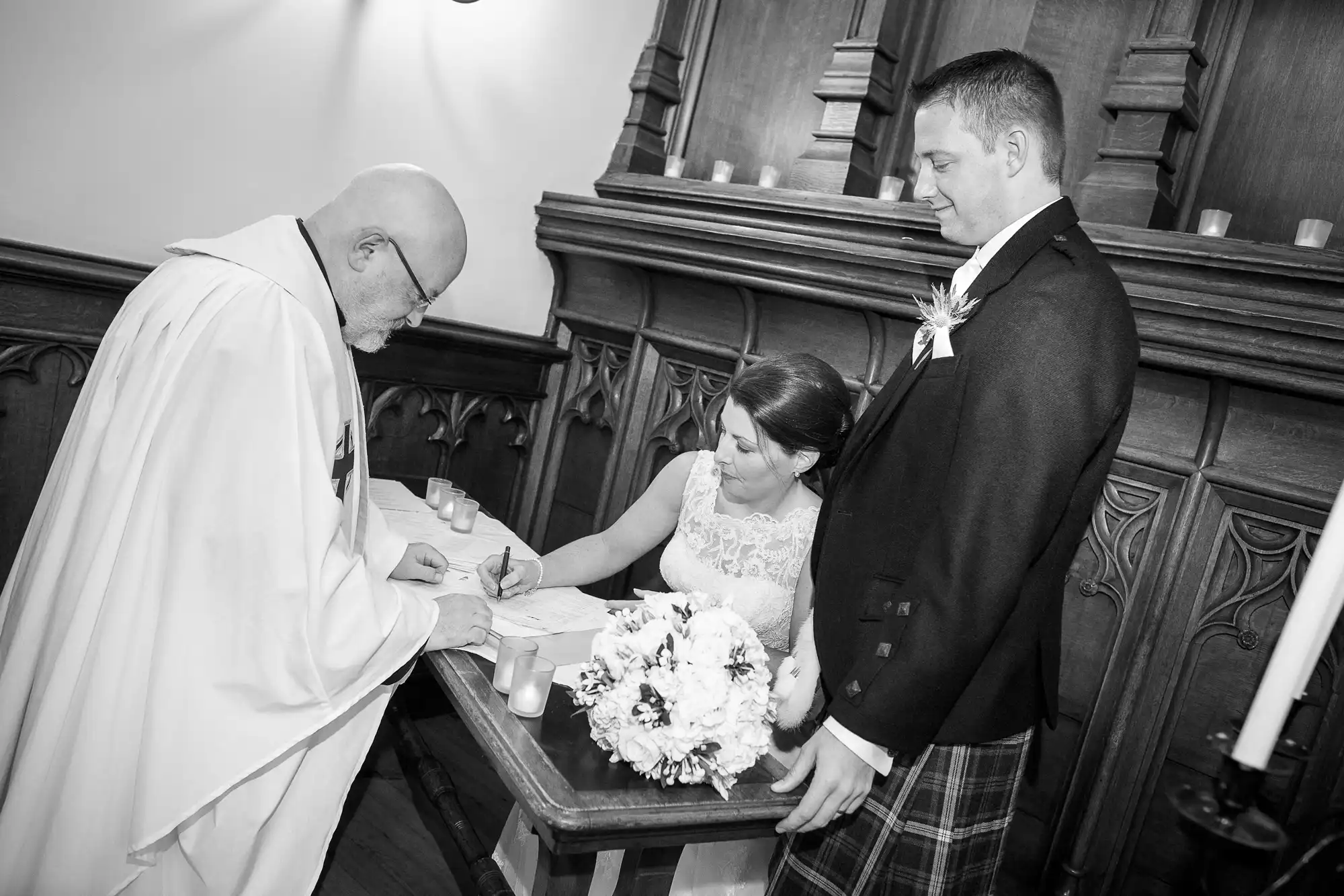 A bride and groom signing their marriage certificate at a church altar, assisted by a priest, in a black and white photo.