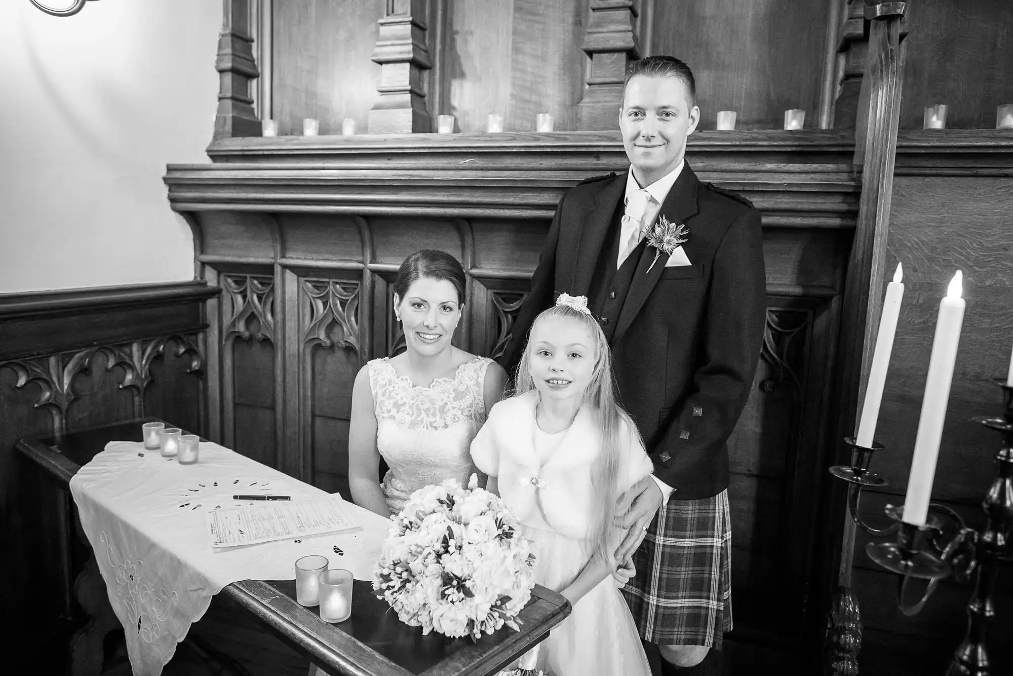 A bride, groom, and young bridesmaid pose beside a signing table with a bouquet and candles in a church ceremony, all in formal attire.