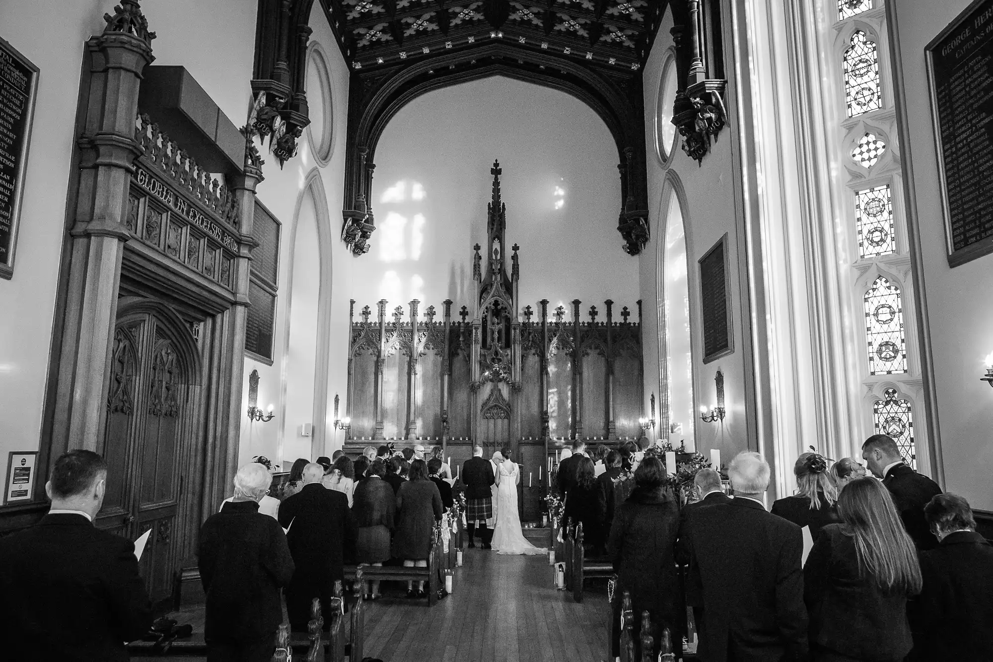 A black and white photo capturing a wedding ceremony inside a traditional church with guests standing as the bride and groom walk down the aisle.