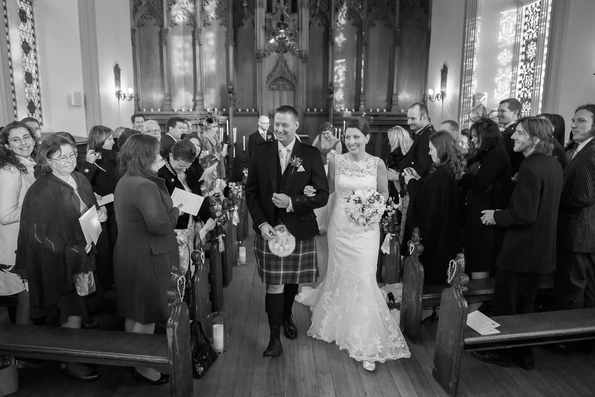 A bride and groom, the groom in a kilt, smiling as they walk down the aisle of a church filled with guests.