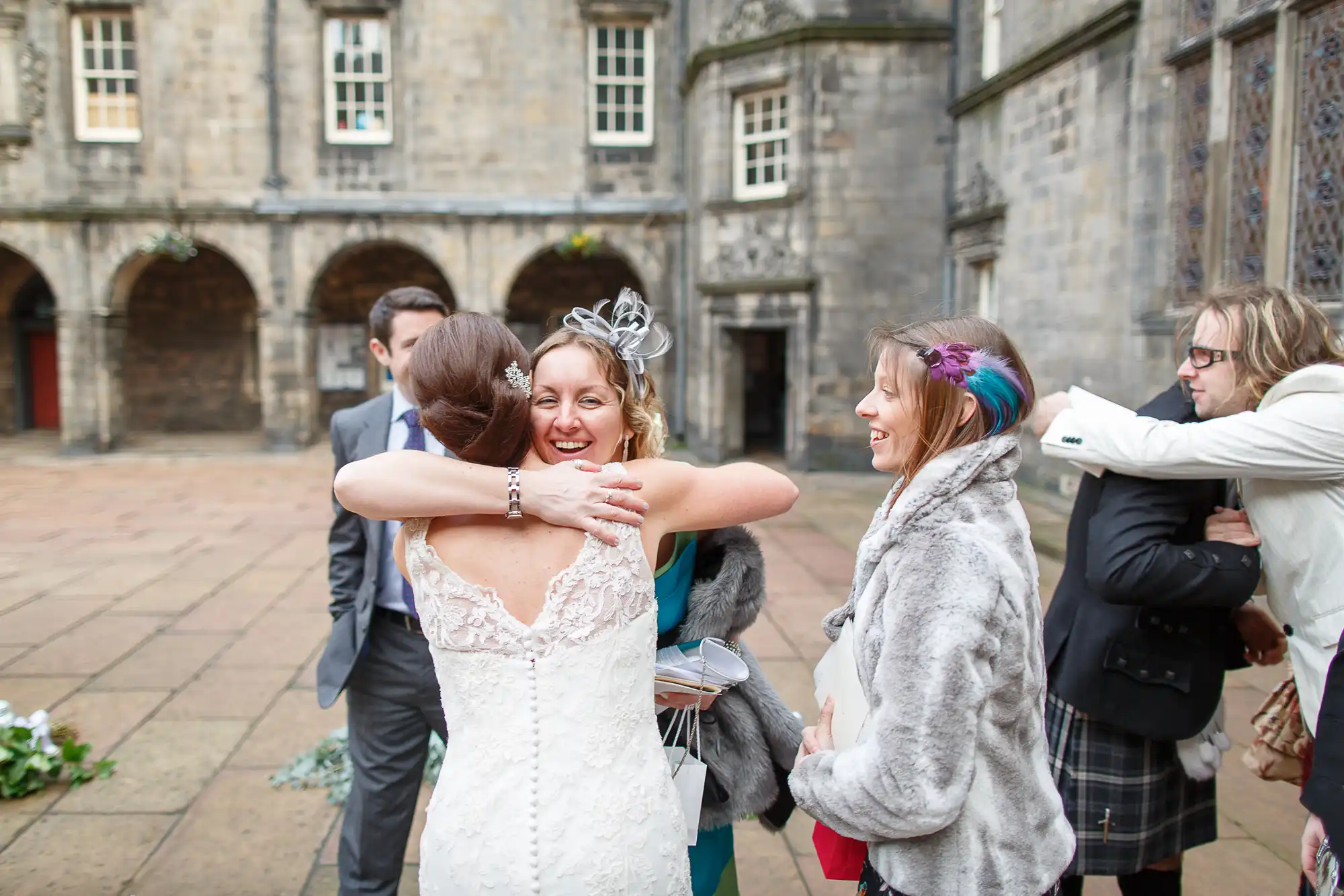 A bride in a white dress embraces a guest in a courtyard, surrounded by other smiling guests during a wedding ceremony.