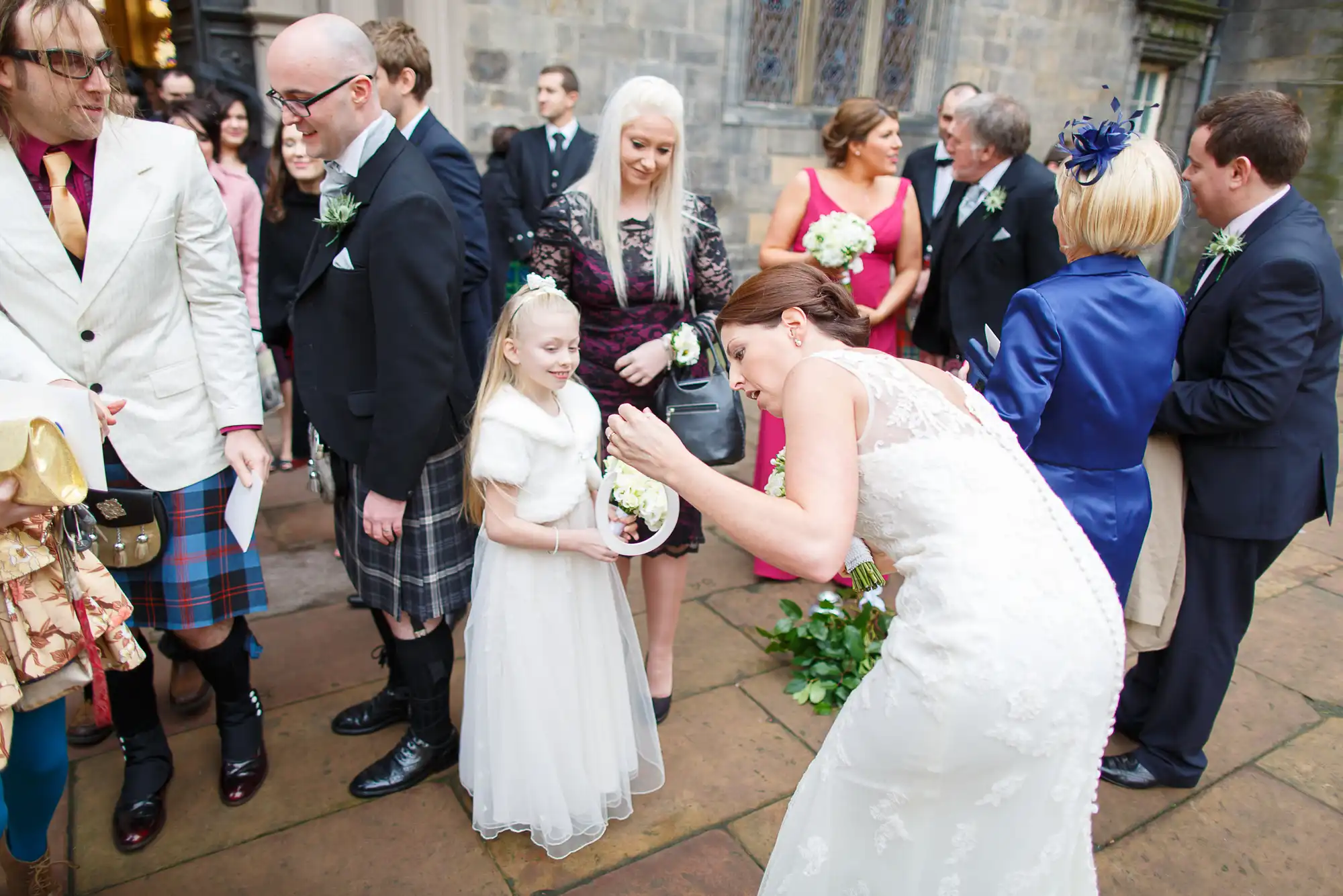A bride in a white dress kneels to speak with a young girl holding a flower basket at a wedding ceremony, surrounded by guests in various formal attires.