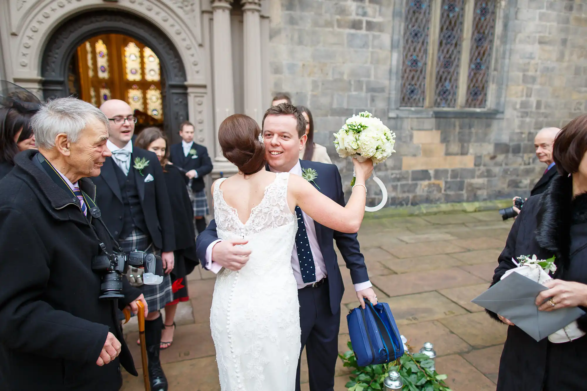 Bride and groom hug outside a church, surrounded by guests, with a photographer capturing the moment.