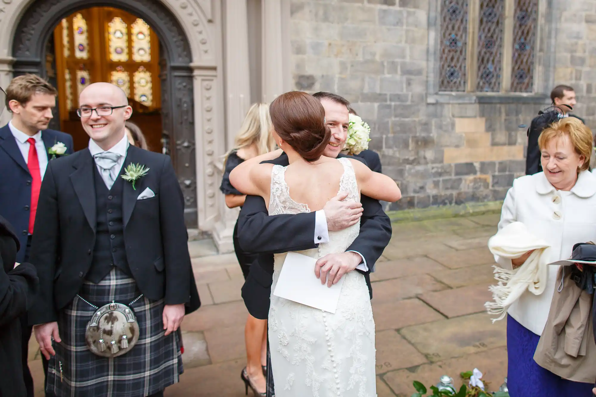A bride in a white lace dress hugs a guest at a wedding ceremony outside a church, as a smiling man in a kilt looks on.