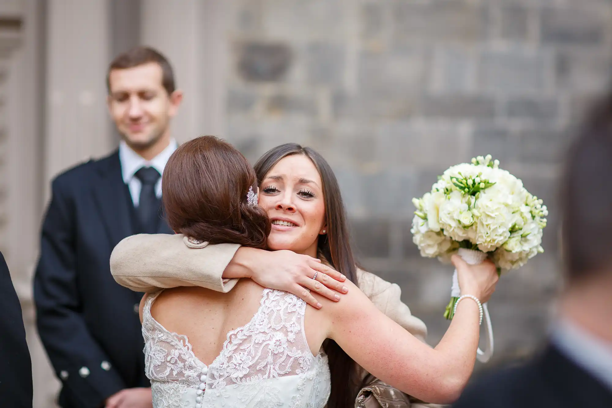 Bride in a lace dress hugs a guest, holding a white bouquet, with a smiling groom in the background.
