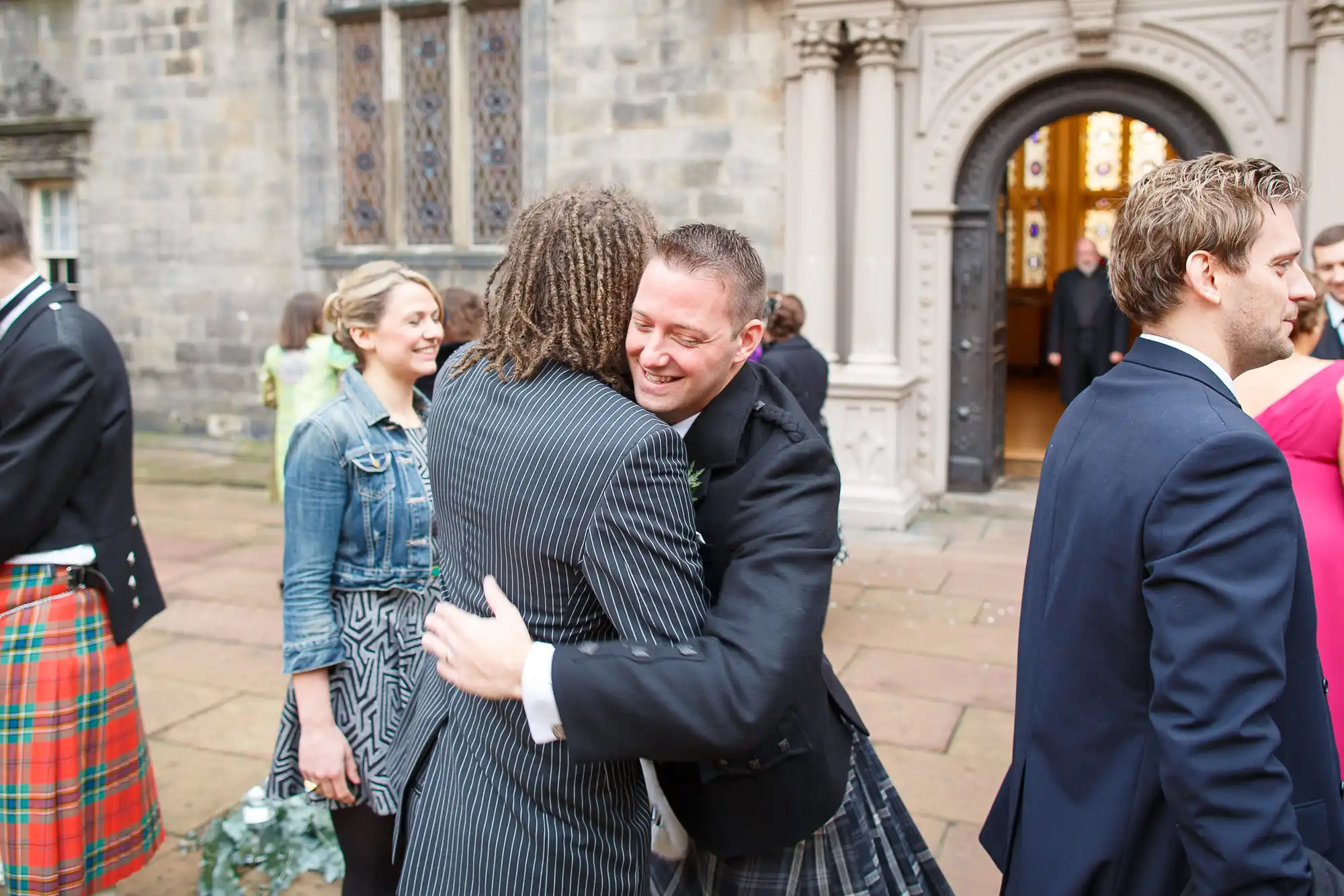 Two men embracing warmly outside a church, one in a striped suit and the other in a formal jacket, with guests in kilts and dresses nearby.