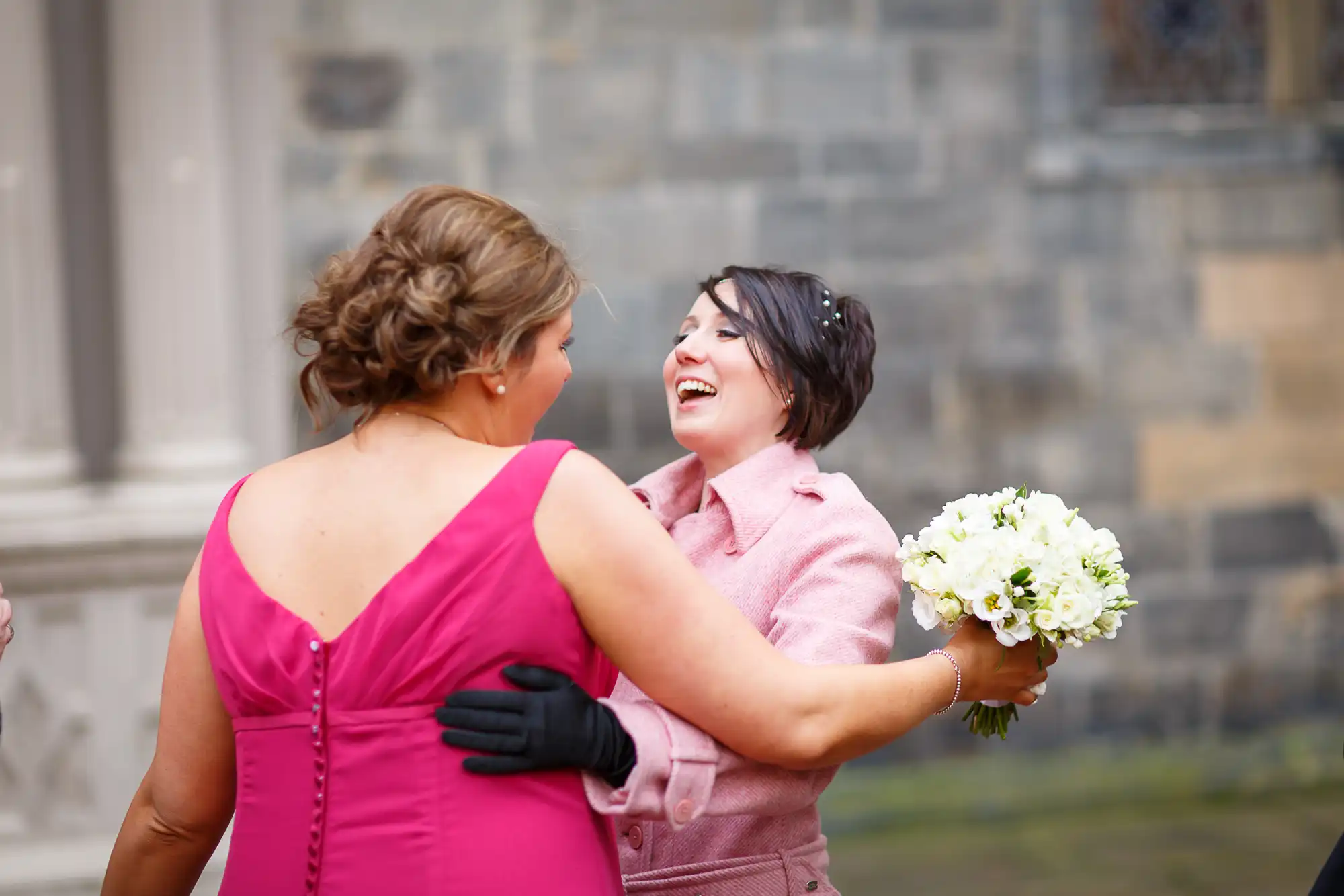 Two women in formal attire hugging and laughing joyously, one holding a bouquet, outside a historical building.