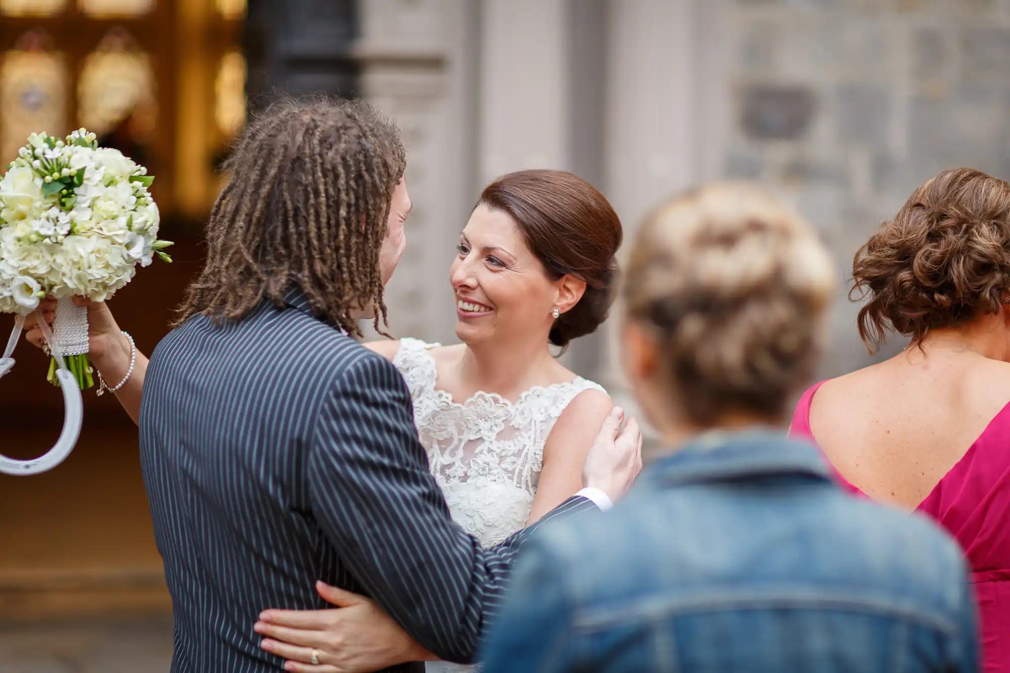 A bride in a lace dress and a groom in a striped suit embracing and smiling at each other, with guests nearby.