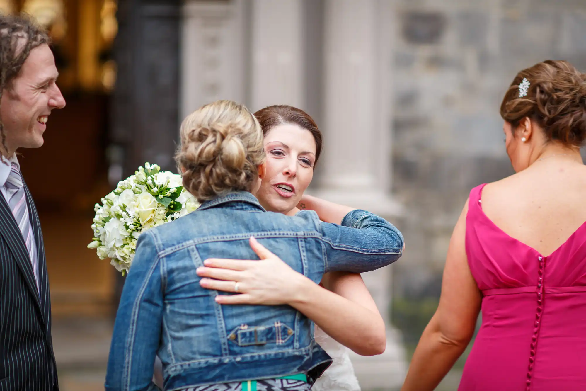 Bride in white dress embraces a woman in a denim jacket, holding a bouquet, while conversing with a smiling man and another woman in pink.