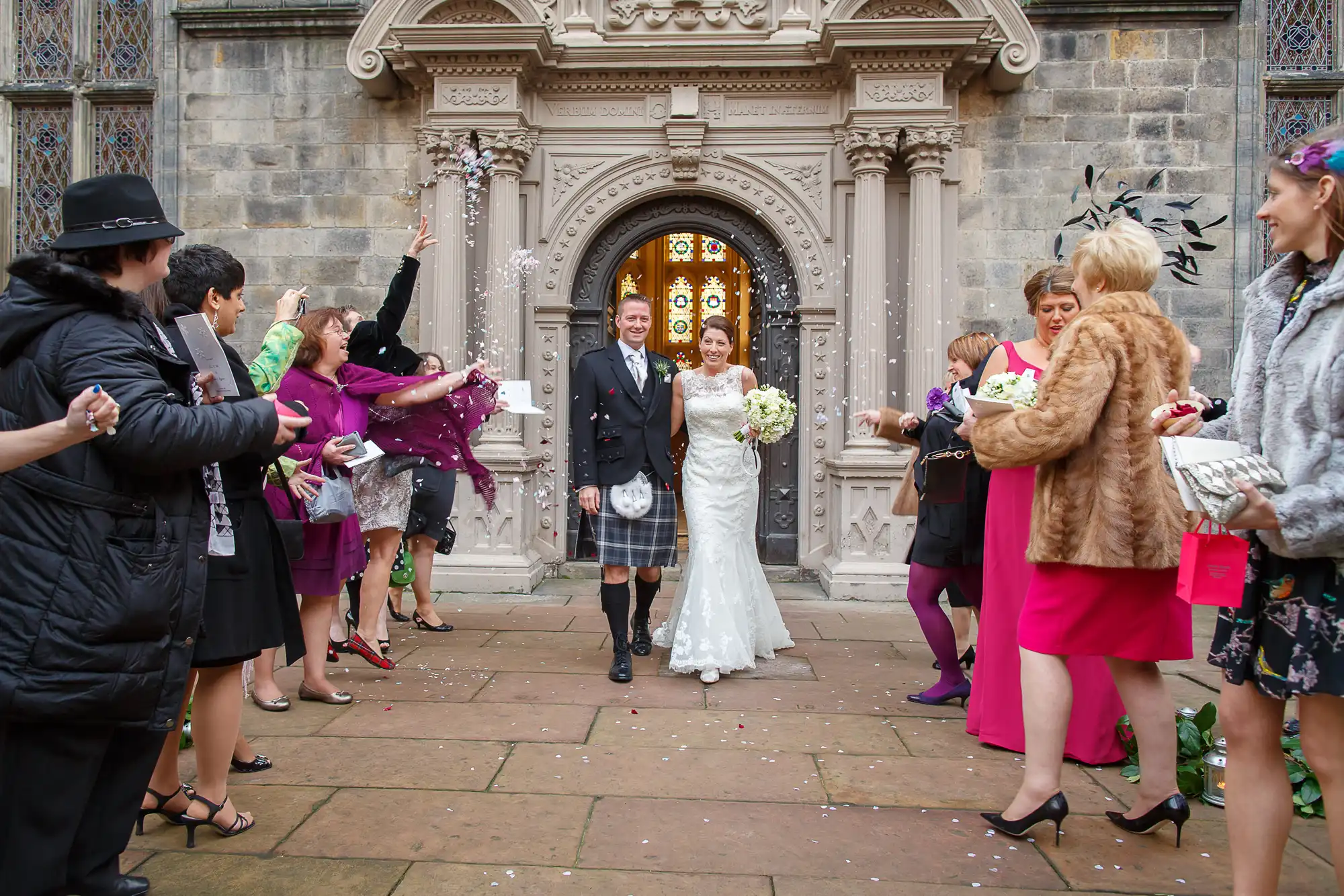 A newlywed couple exits a church as guests throw confetti; the groom wears a kilt, and the bride is in a white dress.