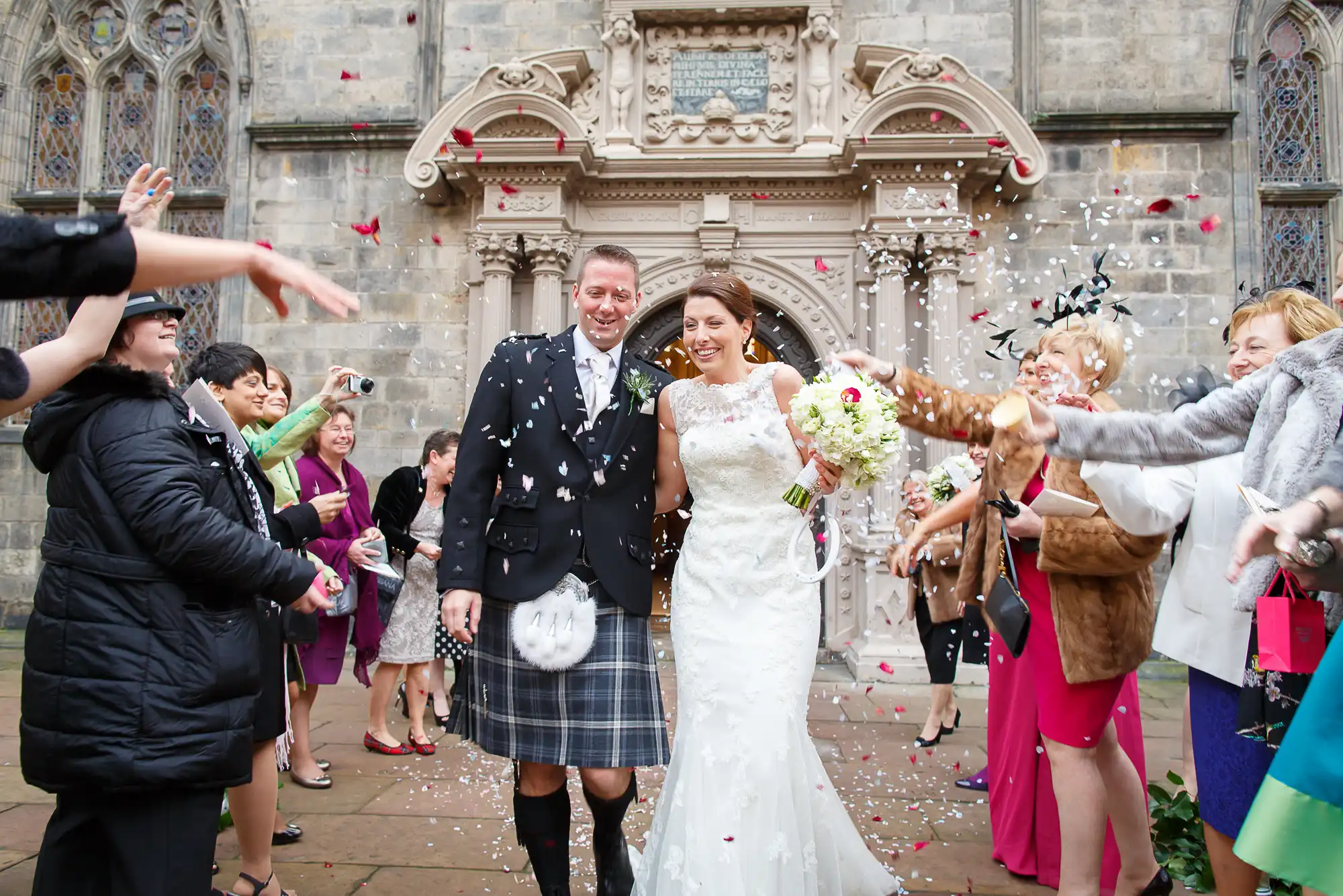 A bride and groom smile as they walk through a crowd throwing confetti outside a church, the groom in a kilt and the bride in a white gown.