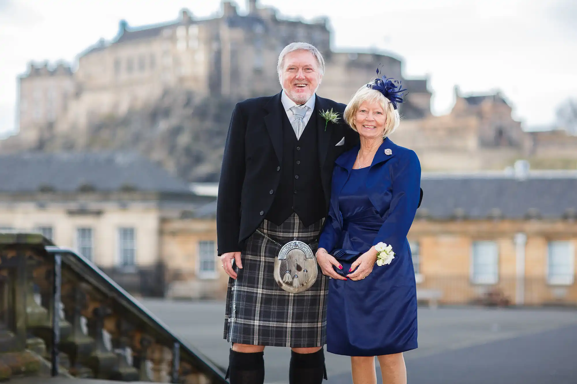 An elderly couple dressed in formal attire with the man in a kilt and the woman in a blue outfit, standing before edinburgh castle.