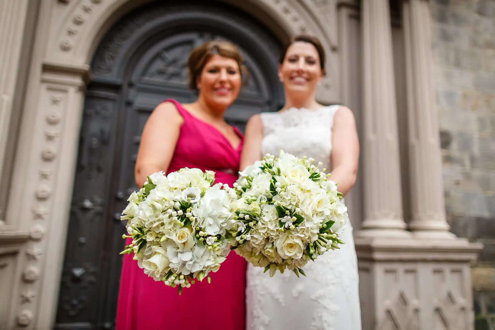 Two women, one in a pink dress and the other in a white wedding gown, holding bouquets and standing in front of an ornate church door.