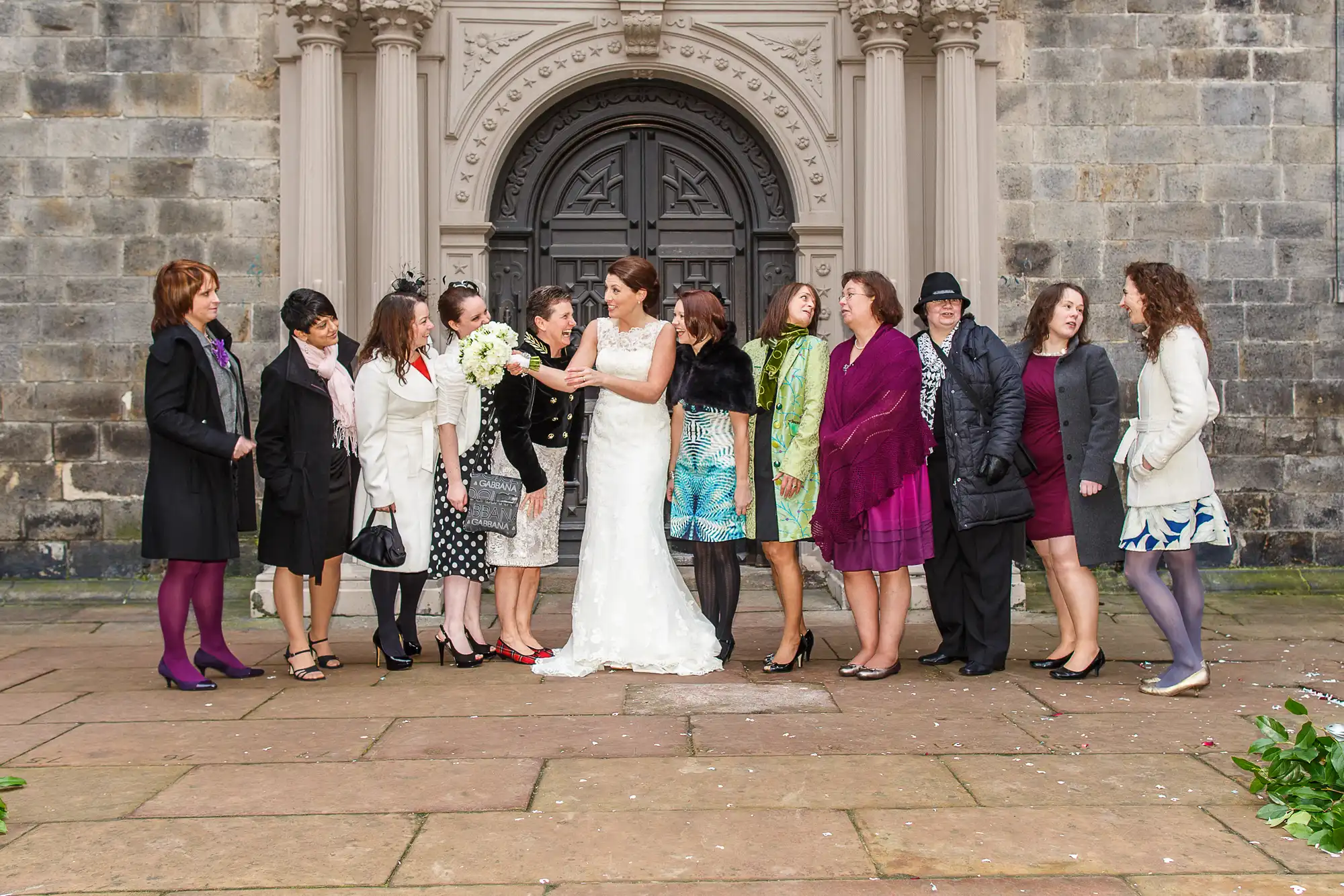 A group of women in various colorful outfits gathers outside a church, interacting cheerfully with a bride who is holding a bouquet.