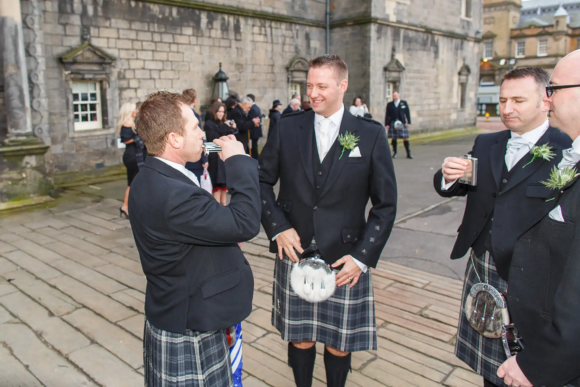 Men in traditional scottish kilts and jackets at a formal gathering, conversing and laughing outside a stone building.