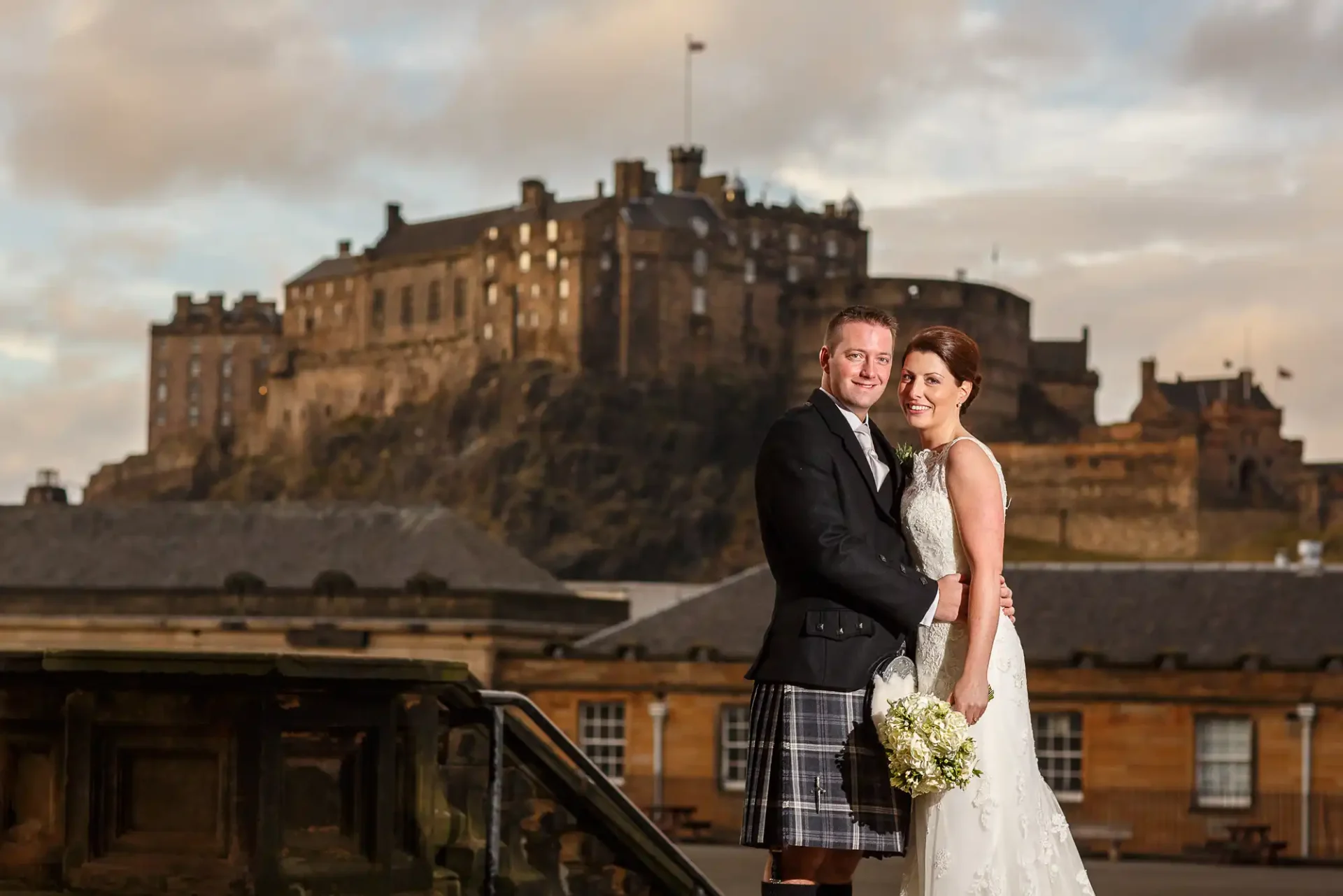 George Heriot's School wedding photographer image of a bride and groom pose together outdoors in front of Edinburgh Castle, with the groom in a kilt and the bride holding a bouquet.