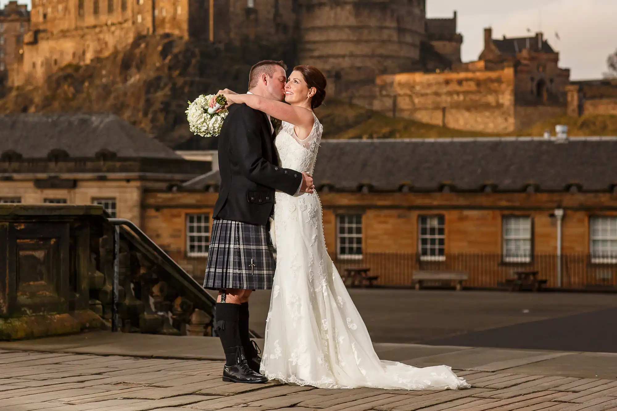 A bride and groom kissing, with the groom in a kilt and the bride in a white gown, in front of a castle on a sunny day.