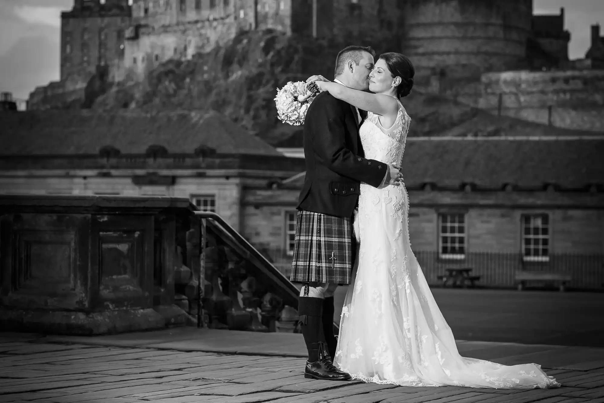 A bride and groom kissing passionately in formal wedding attire, with a historic castle illuminated in the background, all captured in black and white.