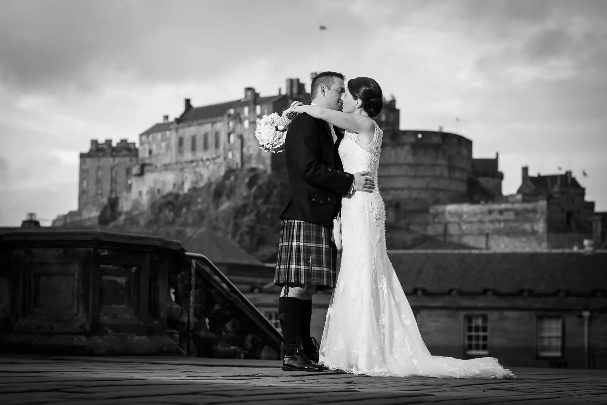 A bride and groom kissing, with the groom dressed in a kilt. edinburgh castle is visible in the background.
