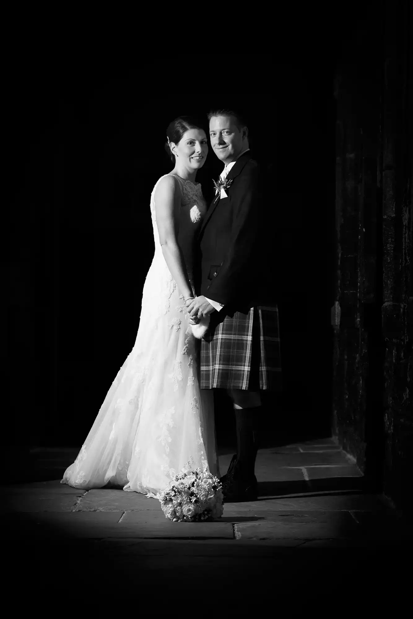 Bride in a white dress and groom in a kilt smiling together in a dimly lit setting with a bouquet on the ground.