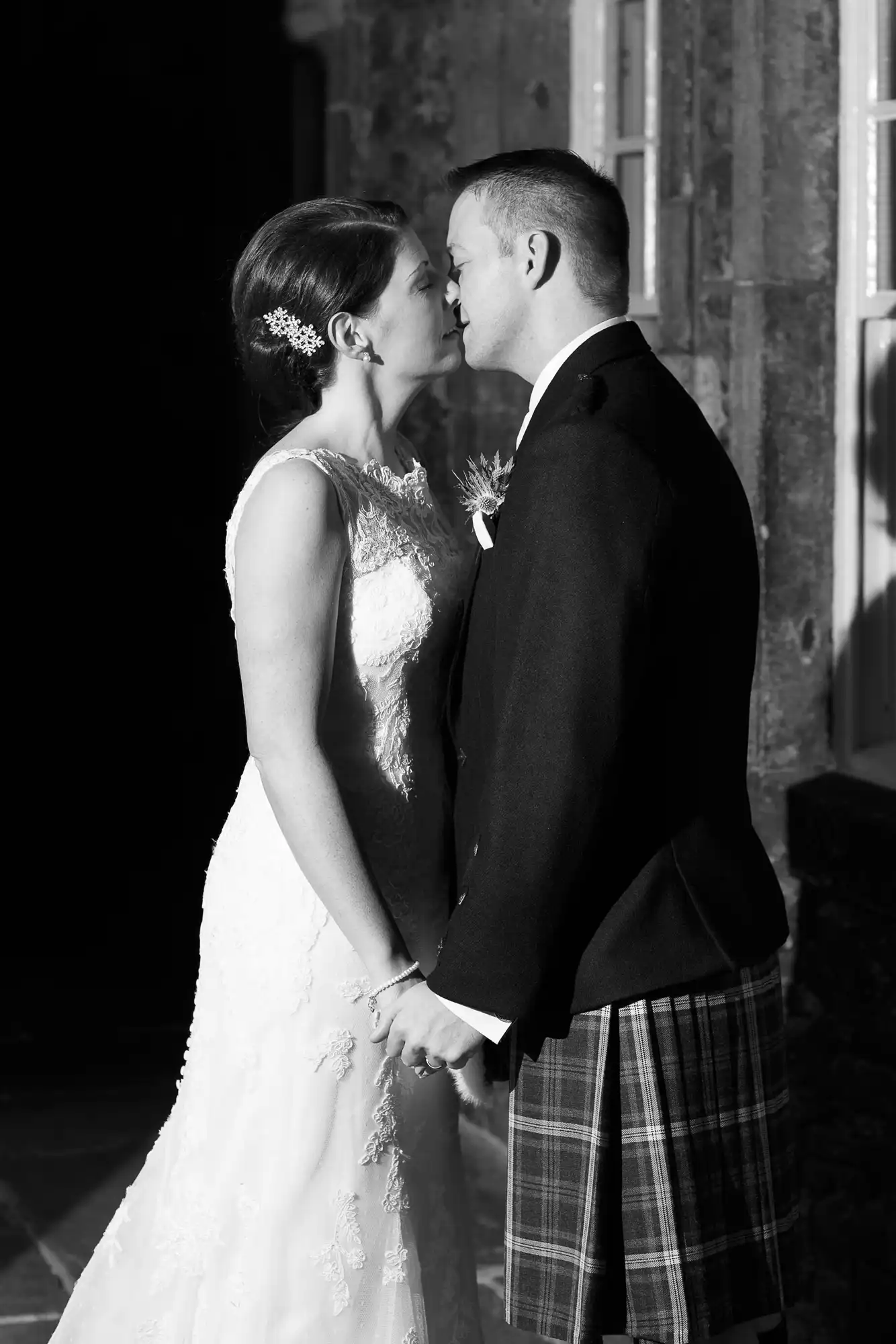 A bride and groom, dressed in formal wedding attire, share a close moment, almost kissing, in dramatic black and white lighting.