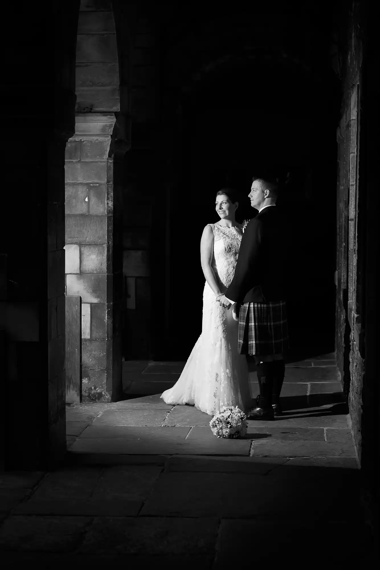 A bride in a white dress and a groom in a kilt stand in a dimly lit archway, illuminated by soft light, exchanging a tender look.