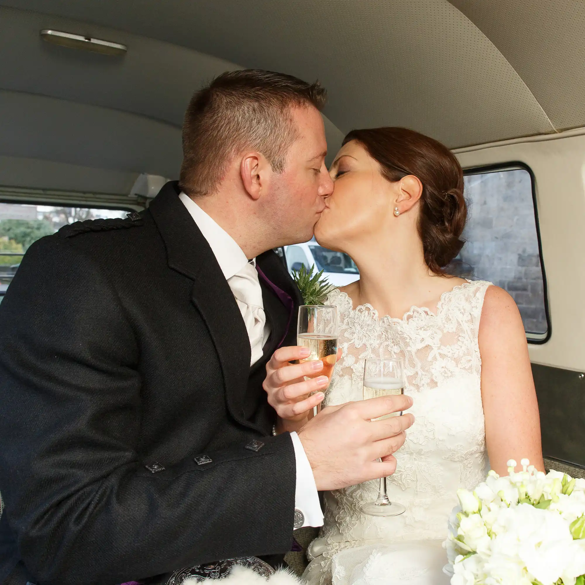 A bride and groom kissing inside a car, each holding a champagne glass, with natural light illuminating the scene.