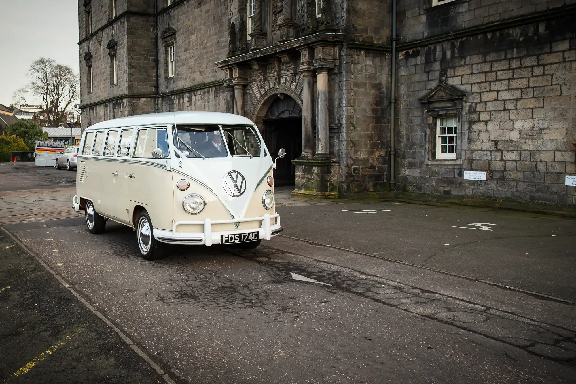 A vintage white volkswagen van parked in front of an old stone building with arched doorways.