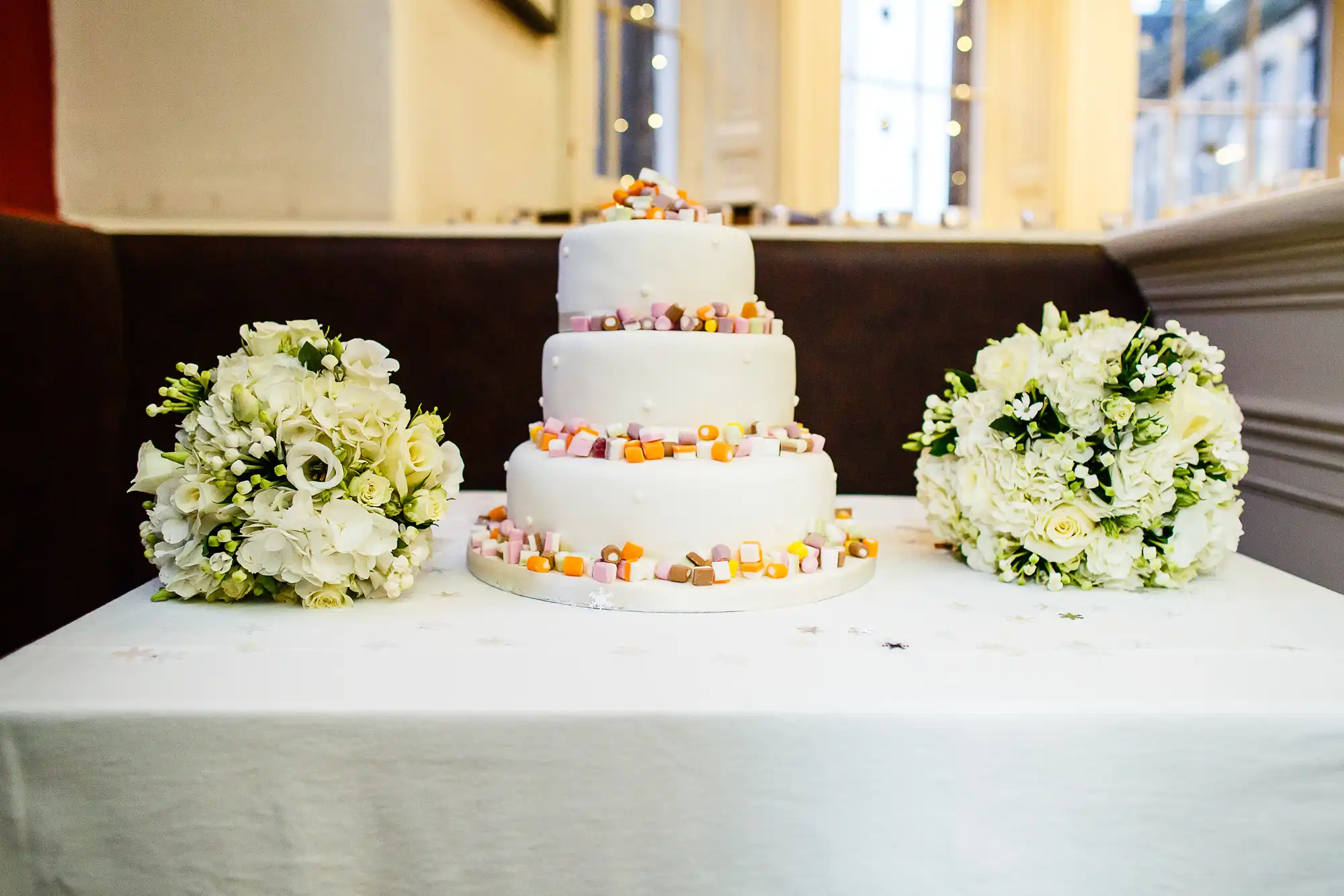 A four-tiered white wedding cake decorated with colorful candy, flanked by two bouquets of white and green flowers on a table.