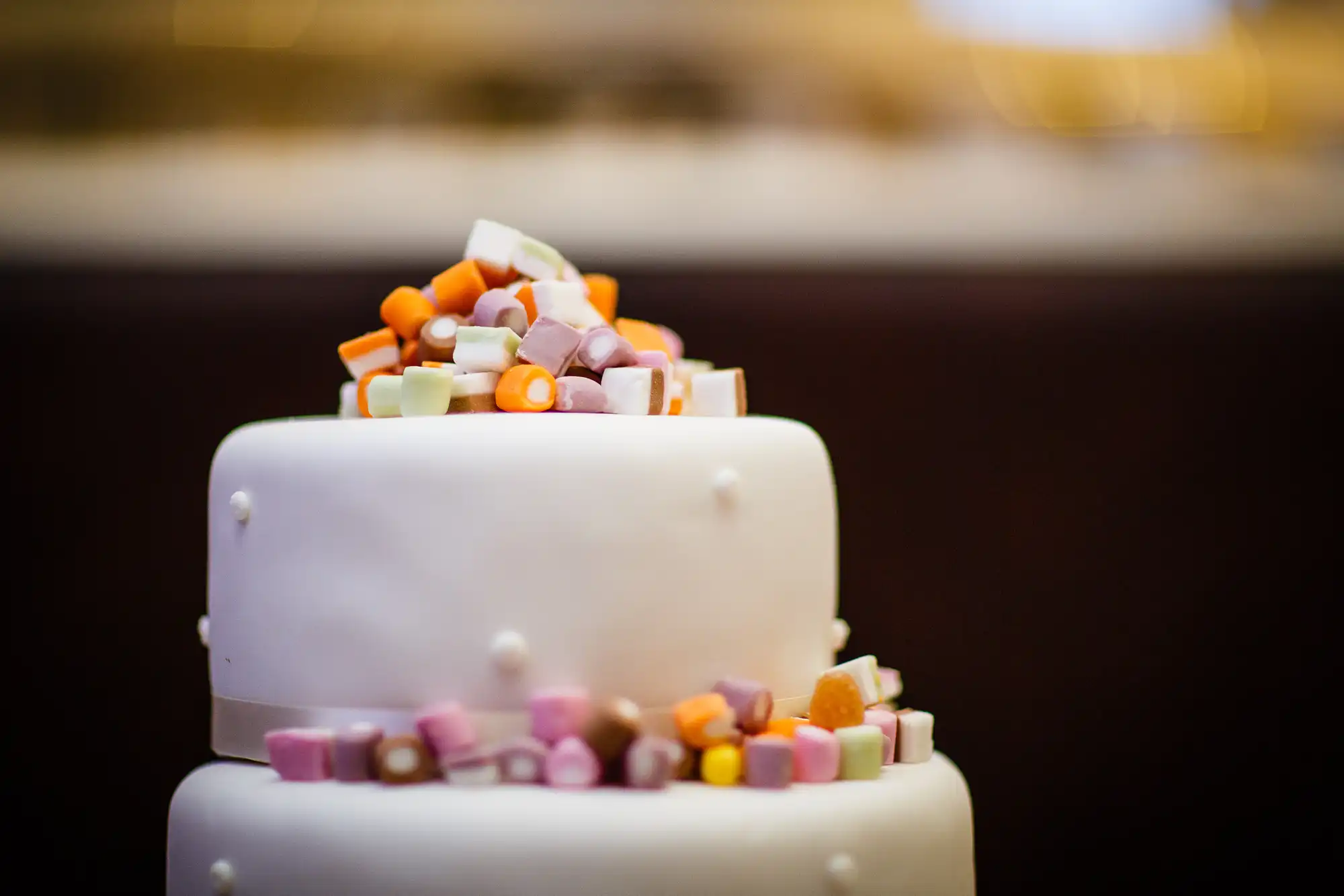 White two-tiered cake decorated with assorted colorful candies on top, set against a softly blurred background.