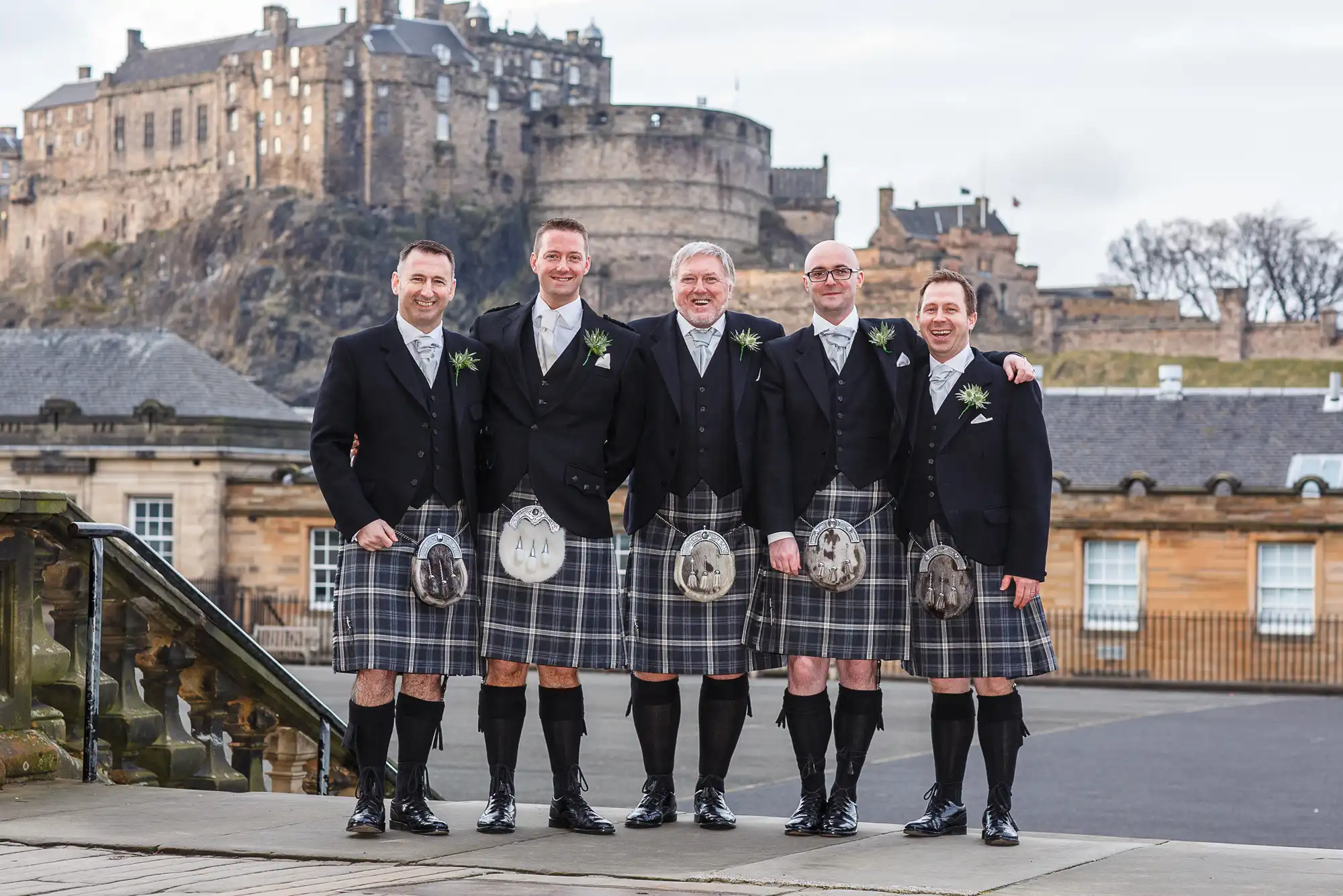 Five men in traditional scottish kilts and formal jackets smiling in front of edinburgh castle.