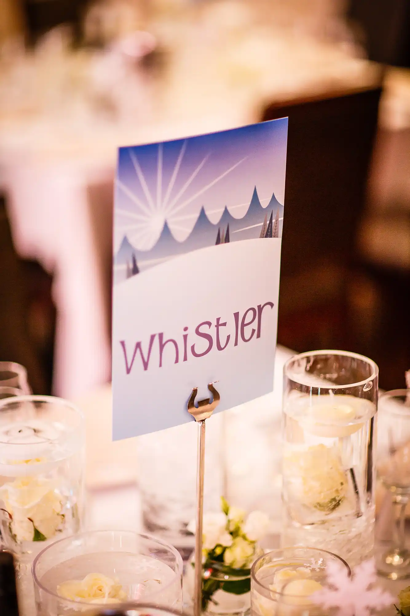 Table sign reading "whistler" with an illustrated mountain backdrop, placed among glasses and floral decorations at an elegant event.