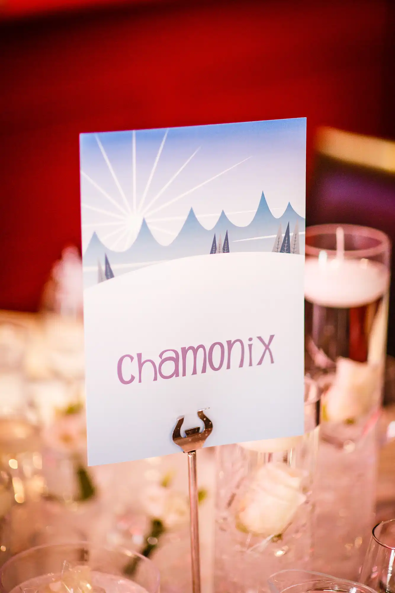 Table marker with "chamonix" printed on it, featuring a graphic of snowy mountain peaks, placed among glassware on a table with a red background.