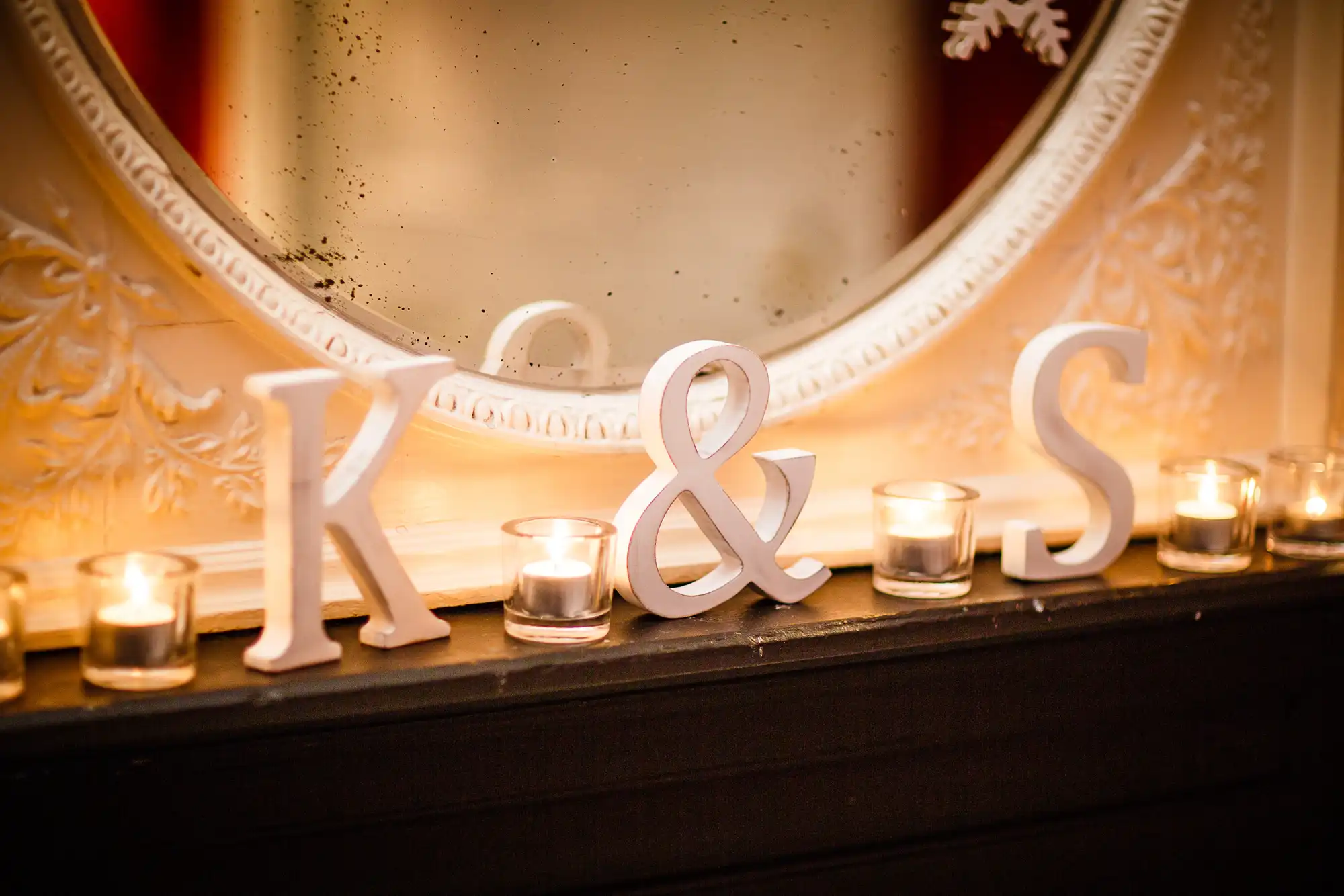 White letters "k & s" on a mantelpiece with lit candles and a decorative mirror in the background.