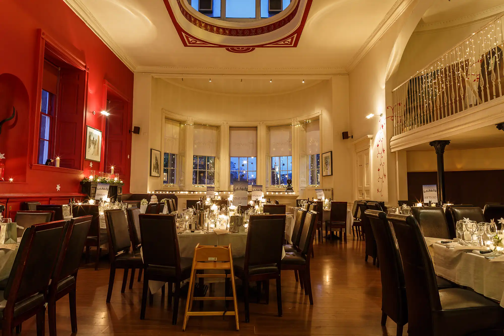Elegant restaurant dining room with red walls, candlelit tables, large windows, and a balcony with decorative railing.