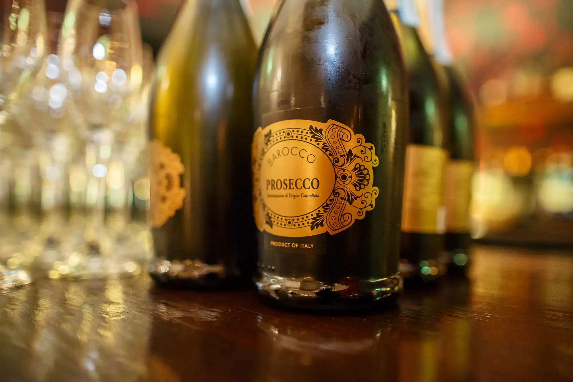 Close-up of prosecco bottles on a bar, focused on the label of one, with empty wine glasses and blurred background.