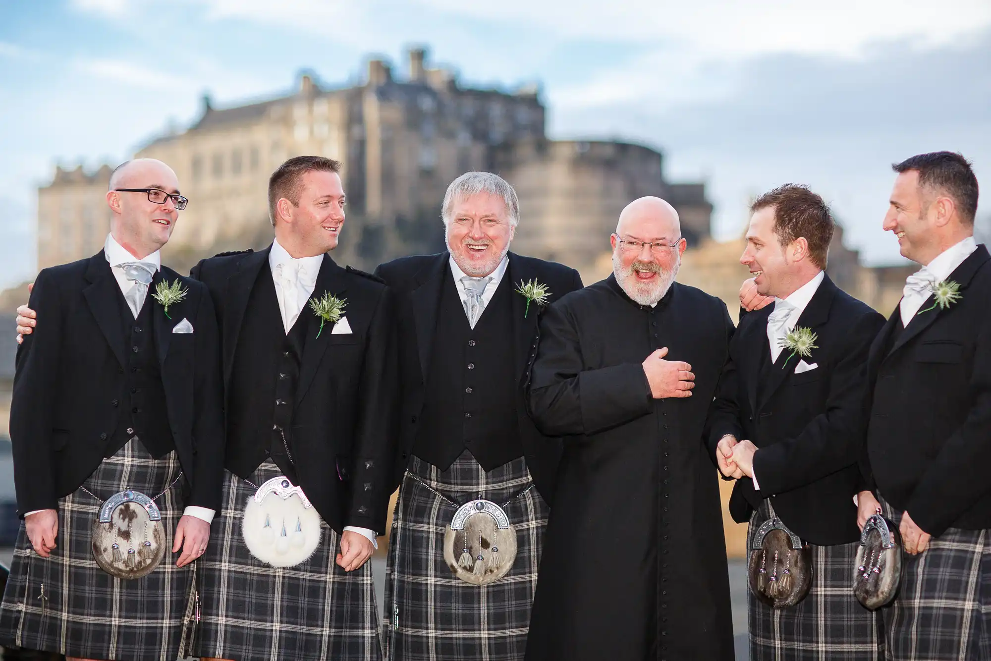 Group of six men in traditional scottish kilts and jackets, smiling and interacting happily, with a castle in the background.