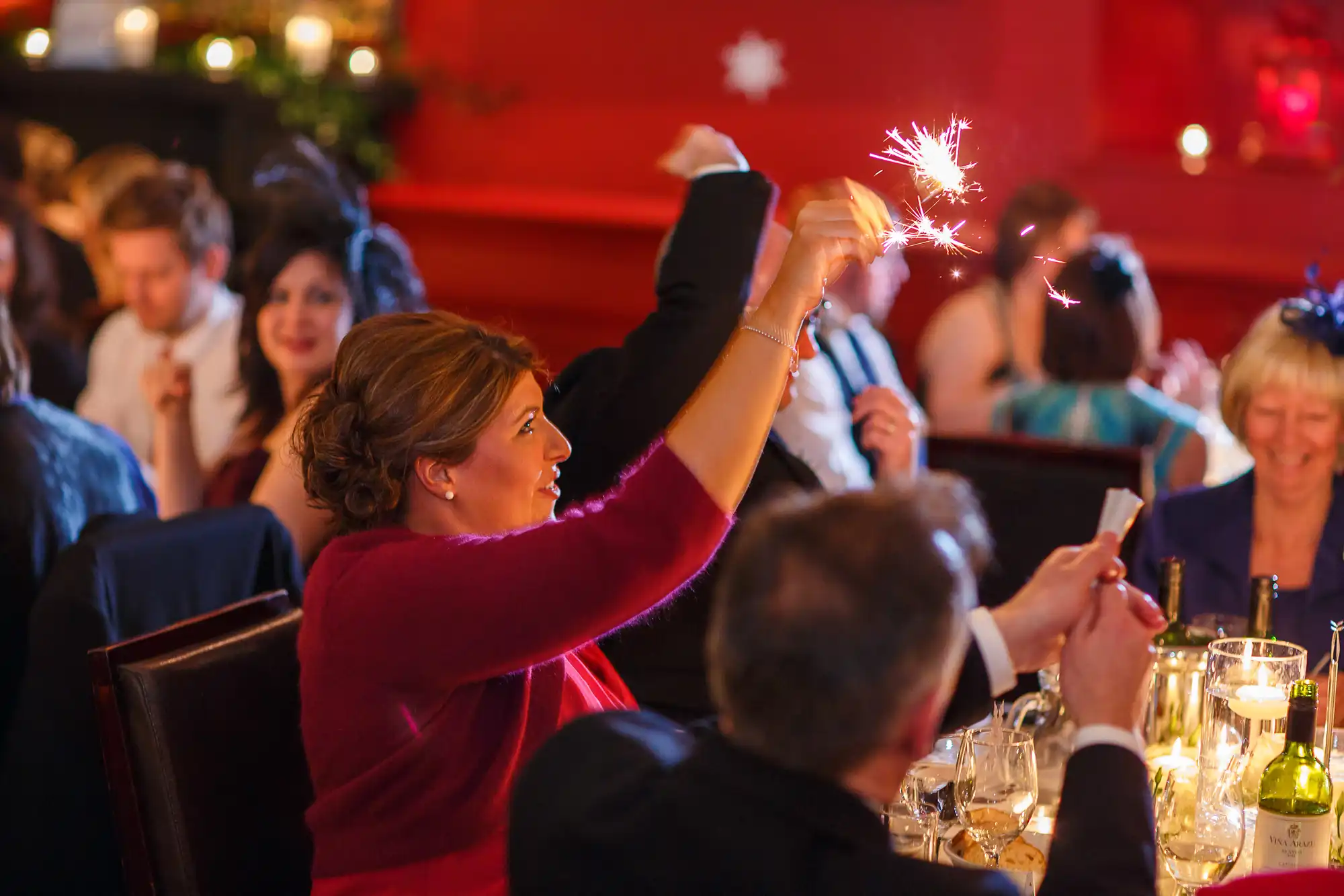 Woman holding a sparkler aloft at a festive dinner party, surrounded by guests in a warmly lit room.